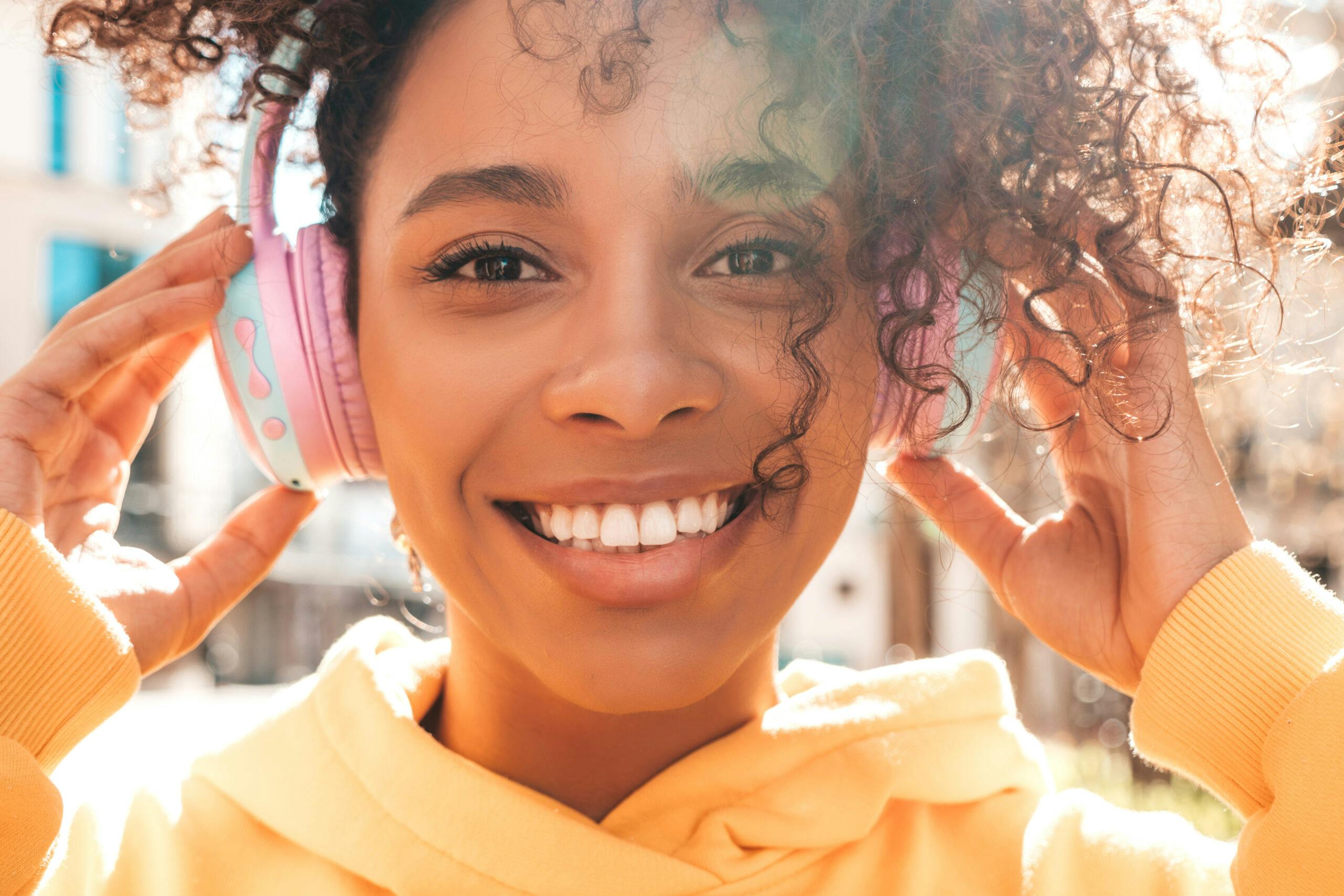 Beautiful black woman with afro curls hairstyle.Smiling model in yellow hoodie.Sexy carefree female enjoying listening music in wireless headphones.Posing on street background at sunset